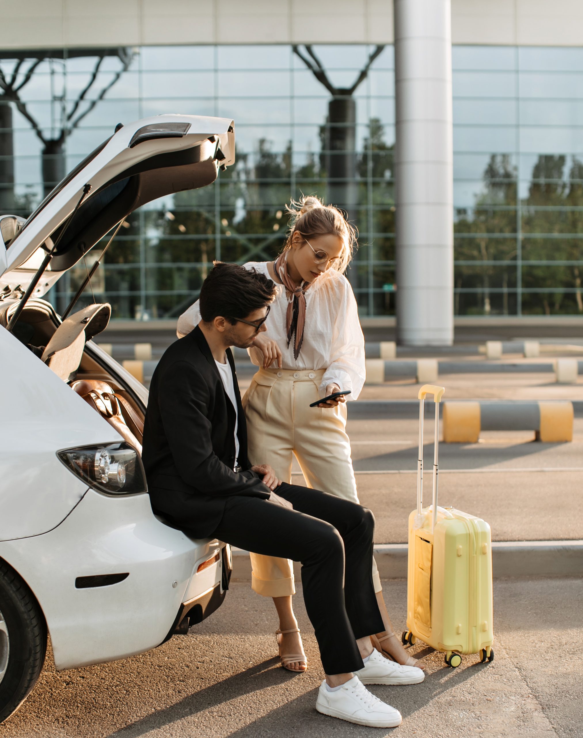 Brunette man in black suit and white t-shirt sits in car trunk. Blonde woman in eyeglasses, white b